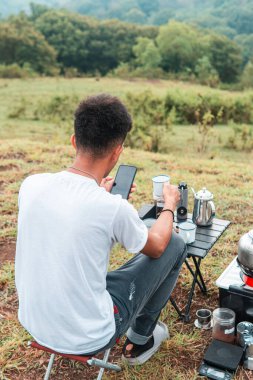 Genç adam, doğada dinlendirici bir kamp gezisi sırasında akıllı telefonunu kullanarak bir fincan kahvenin tadını çıkarıyor.