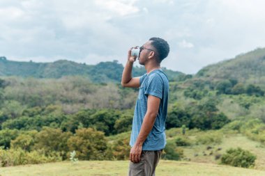 Endonezyalı genç adam kahve içiyor. Etrafı nefes kesici tropik dağ manzaralarıyla çevrili. Doğada huzurlu bir anın tadını çıkarıyor.