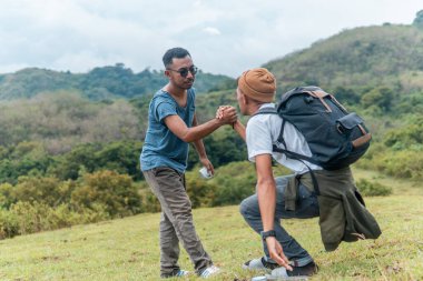 Güneydoğu Asyalı bir adam arkadaşının çimenli bir tepeye çıkmasına yardım ediyor. Destek, birlik, takım çalışması ve açık hava dostluğunun sembolü. Motivasyon, nezaket ve toplum kavramları için harika..