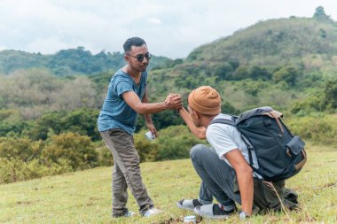 Güneydoğu Asyalı bir adam arkadaşının çimenli bir tepeye çıkmasına yardım ediyor. Destek, birlik, takım çalışması ve açık hava dostluğunun sembolü. Motivasyon, nezaket ve toplum kavramları için harika..