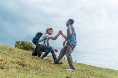 Güneydoğu Asyalı bir adam arkadaşının çimenli bir tepeye çıkmasına yardım ediyor. Destek, birlik, takım çalışması ve açık hava dostluğunun sembolü. Motivasyon, nezaket ve toplum kavramları için harika..