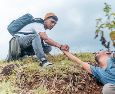 Bir adamın dağa tırmanmasına yardım etmek takım çalışmasını, desteği ve açık hava macerasını sembolize eder. Bulutlu gökyüzünün altında doğa yürüyüşü sırasında çekilen.
