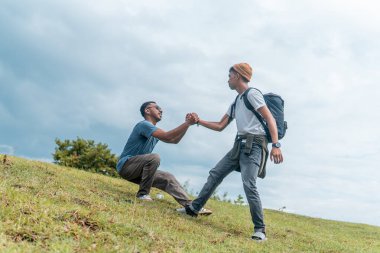Güneydoğu Asyalı bir adam arkadaşının çimenli bir tepeye çıkmasına yardım ediyor. Destek, birlik, takım çalışması ve açık hava dostluğunun sembolü. Motivasyon, nezaket ve toplum kavramları için harika..