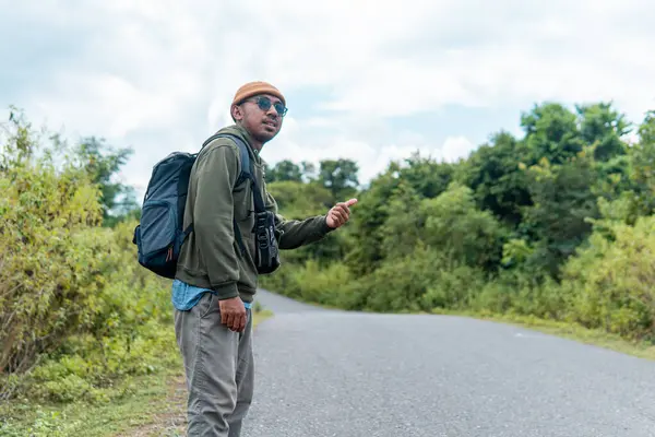 Sırt çantalı bir adam kırsal bir yolda otostop çekiyor. Yeşillik ve bulutlu gökyüzü ile çevrelenmiş bu görüntü tek başına seyahat etmenin, özgürlüğün ve vahşi doğada maceranın ruhunu yakalıyor..