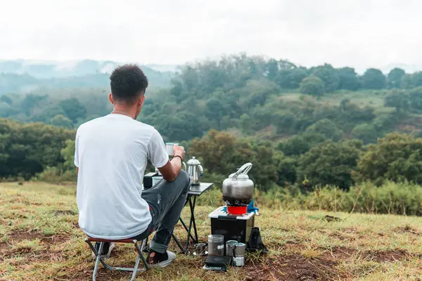 Genç adam doğada kamp yaparken rahatlıyor ve kahve içiyor. Yeşil tepelerin huzurlu manzarasının tadını çıkarıyor.