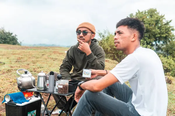 İki Endonezyalı adam kahve hazırlarken rahat bir ortamda birlikte gülüyorlar. Rahat bir kamp, kaynaşma ve çimenli bir arazide taşınabilir kahve takımlarıyla doğanın tadını çıkarma anı..