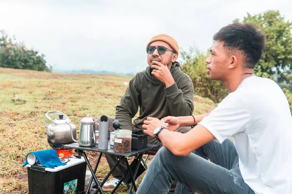İki Endonezyalı adam kahve hazırlarken rahat bir ortamda birlikte gülüyorlar. Rahat bir kamp, kaynaşma ve çimenli bir arazide taşınabilir kahve takımlarıyla doğanın tadını çıkarma anı..