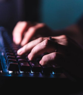 Karanlık bir odada LED arka ışığı olan mekanik bir klavyede yazı yazan yakın çekim elleri. Sığ alan derinliği ve yumuşak ışıklandırma bir teknoloji ve üretkenlik ortamı yaratır..