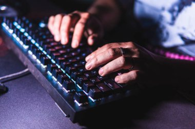 Karanlık bir odada LED arka ışığı olan mekanik bir klavyede yazı yazan yakın çekim elleri. Sığ alan derinliği ve yumuşak ışıklandırma bir teknoloji ve üretkenlik ortamı yaratır..