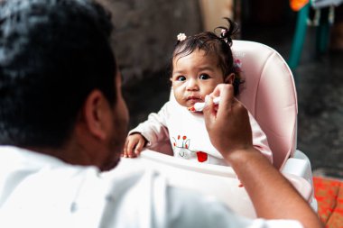 Bir baba evde yemek saatinde bebeğini kaşıkla besliyor. Bebek, yüksek bir sandalyede oturup aile bağlarını ve bakımını gösterirken meraklı ve sevimli görünüyor..