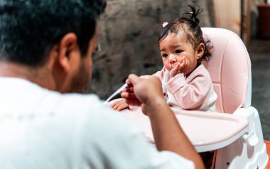 Bir baba evde yemek saatinde bebeğini kaşıkla besliyor. Bebek, yüksek bir sandalyede oturup aile bağlarını ve bakımını gösterirken meraklı ve sevimli görünüyor..