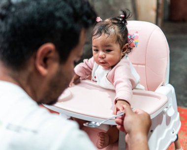 Bir baba evde yemek saatinde bebeğini kaşıkla besliyor. Bebek, yüksek bir sandalyede oturup aile bağlarını ve bakımını gösterirken meraklı ve sevimli görünüyor..