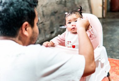 Bir baba evde yemek saatinde bebeğini kaşıkla besliyor. Bebek, yüksek bir sandalyede oturup aile bağlarını ve bakımını gösterirken meraklı ve sevimli görünüyor..