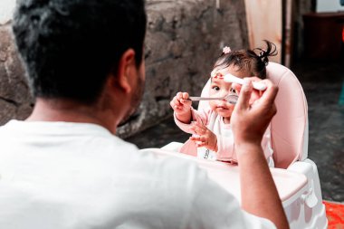 Bir baba evde yemek saatinde bebeğini kaşıkla besliyor. Bebek, yüksek bir sandalyede oturup aile bağlarını ve bakımını gösterirken meraklı ve sevimli görünüyor..