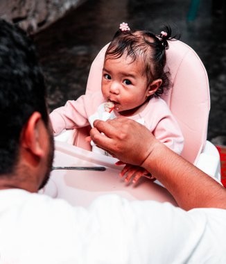 Bir baba evde yemek saatinde bebeğini kaşıkla besliyor. Bebek, yüksek bir sandalyede oturup aile bağlarını ve bakımını gösterirken meraklı ve sevimli görünüyor..