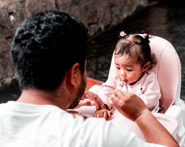 Bir baba evde yemek saatinde bebeğini kaşıkla besliyor. Bebek, yüksek bir sandalyede oturup aile bağlarını ve bakımını gösterirken meraklı ve sevimli görünüyor..