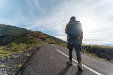 Sırt çantalı yalnız bir yürüyüşçü güneşin doğduğu gökyüzünün altında boş bir dağ yolunda yürüyor. Keşif ve azmi simgeliyor..