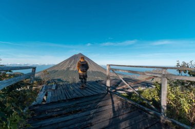 Sarı ceketli ve sırt çantalı Asyalı bir yürüyüşçü Endonezya 'nın Doğu Nusa Tenggara kentindeki manzaralı ahşap bir perspektiften Inerie Dağı' na hayran. Macera, doğa ve seyahat kavramları için mükemmel..