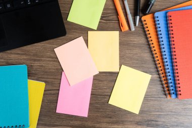 Tahta bir masadaki boş yapışkan notların üst görüntüsü açık bir dizüstü bilgisayar, renkli dosyalar ve kalemler. Ofis, planlama, üretkenlik ve yaratıcı çalışma alanı kavramları için mükemmel..