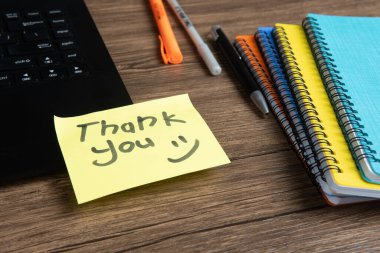 Elle yazılmış sarı yapışkan not. Açık bir dizüstü bilgisayara yerleştirilmiş. Etrafı renkli ciltler, kalemler ve ahşap bir masadaki diğer ofis malzemeleriyle çevrili. Minnettarlık ve ofis kavramları için mükemmel..