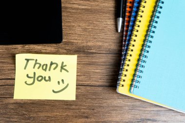 Elle yazılmış sarı yapışkan not. Açık bir dizüstü bilgisayara yerleştirilmiş. Etrafı renkli ciltler, kalemler ve ahşap bir masadaki diğer ofis malzemeleriyle çevrili. Minnettarlık ve ofis kavramları için mükemmel..