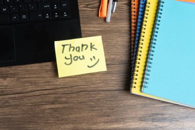 Elle yazılmış sarı yapışkan not. Açık bir dizüstü bilgisayara yerleştirilmiş. Etrafı renkli ciltler, kalemler ve ahşap bir masadaki diğer ofis malzemeleriyle çevrili. Minnettarlık ve ofis kavramları için mükemmel..