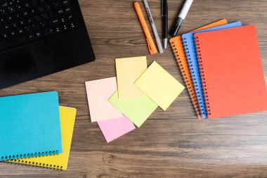 Tahta bir masadaki boş yapışkan notların üst görüntüsü açık bir dizüstü bilgisayar, renkli dosyalar ve kalemler. Ofis, planlama, üretkenlik ve yaratıcı çalışma alanı kavramları için mükemmel..