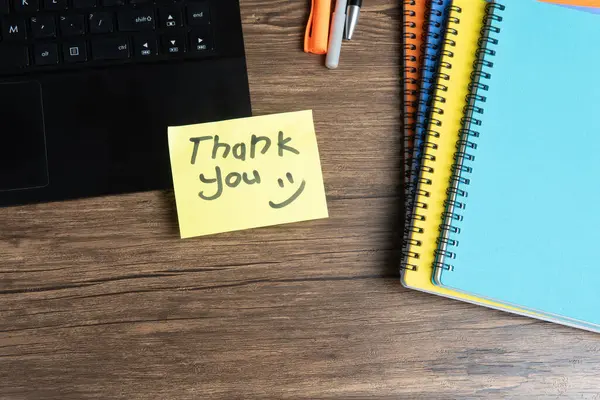 Elle yazılmış sarı yapışkan not. Açık bir dizüstü bilgisayara yerleştirilmiş. Etrafı renkli ciltler, kalemler ve ahşap bir masadaki diğer ofis malzemeleriyle çevrili. Minnettarlık ve ofis kavramları için mükemmel..