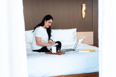 Yatakta bacak bacak üstüne atmış bir Asyalı kadın, dizüstü bilgisayarı ve kahve fincanıyla yakınlarda bir not defterine yazıyor, rahat bir yatak odasında uzaktan çalışmayı, planlamayı ve yaratıcı düşünmeyi temsil ediyor..