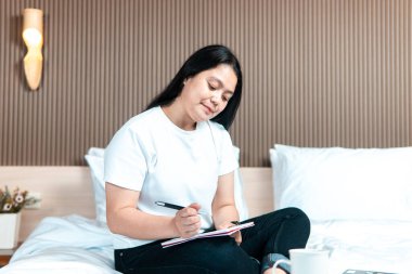 Yatakta bacak bacak üstüne atmış bir Asyalı kadın, dizüstü bilgisayarı ve kahve fincanıyla yakınlarda bir not defterine yazıyor, rahat bir yatak odasında uzaktan çalışmayı, planlamayı ve yaratıcı düşünmeyi temsil ediyor..