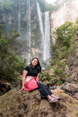 Arkasında tropik bir şelale olan bir kayanın üzerinde oturan Endonezyalı kadın. Serüven, rahatlama, eko turizm, özgürlük, doğa yolculuğu ve bereketli yağmur ormanlarında keşif kavramı.