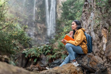 Sarı ceketli ve sırt çantalı Endonezyalı bir kadın bir şelalenin yakınındaki kayaların üzerinde oturuyor, elinde bir defter ve kalem, doğada günlük tutuyor. Farkındalık, seyahat ve ilham kavramı.
