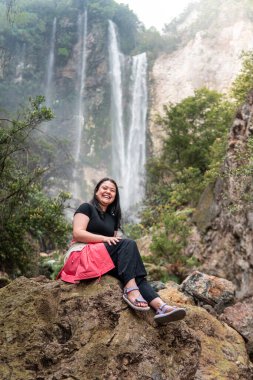 Arkasında tropik bir şelale olan bir kayanın üzerinde oturan Endonezyalı kadın. Serüven, rahatlama, eko turizm, özgürlük, doğa yolculuğu ve bereketli yağmur ormanlarında keşif kavramı.