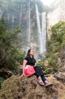 Arkasında tropik bir şelale olan bir kayanın üzerinde oturan Endonezyalı kadın. Serüven, rahatlama, eko turizm, özgürlük, doğa yolculuğu ve bereketli yağmur ormanlarında keşif kavramı.