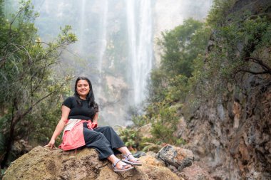 Arkasında tropik bir şelale olan bir kayanın üzerinde oturan Endonezyalı kadın. Serüven, rahatlama, eko turizm, özgürlük, doğa yolculuğu ve bereketli yağmur ormanlarında keşif kavramı.