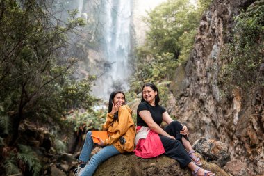İki Endonezyalı kadın, Endonezya 'da tropik bir şelalenin önünde gülümseyerek arkadaşlığın tadını çıkarıyorlar. Seyahat, arkadaşlık, macera ve doğa yaşam tarzı temaları için mükemmel..