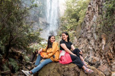 İki Endonezyalı kadın, Endonezya 'da tropik bir şelalenin önünde gülümseyerek arkadaşlığın tadını çıkarıyorlar. Seyahat, arkadaşlık, macera ve doğa yaşam tarzı temaları için mükemmel..