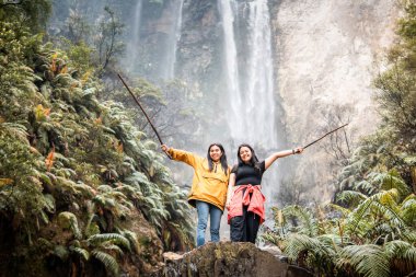 İki Endonezyalı kadın, Endonezya 'da tropik bir şelalenin önünde gülümseyerek arkadaşlığın tadını çıkarıyorlar. Seyahat, arkadaşlık, macera ve doğa yaşam tarzı temaları için mükemmel..