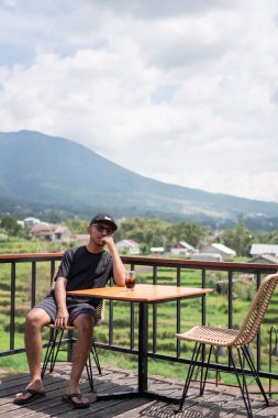 Siyah t-shirt ve şapka giyen Asyalı bir adam, Endonezya 'nın manzaralı dağ ve pirinç tarlalarının tadını çıkarırken terasta oturup kahve içiyor. Rahatlatıcı yaşam tarzı ve seyahat kavramı.