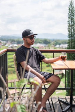 Siyah t-shirt ve şapka giyen Asyalı bir adam, Endonezya 'nın manzaralı dağ ve pirinç tarlalarının tadını çıkarırken terasta oturup kahve içiyor. Rahatlatıcı yaşam tarzı ve seyahat kavramı.