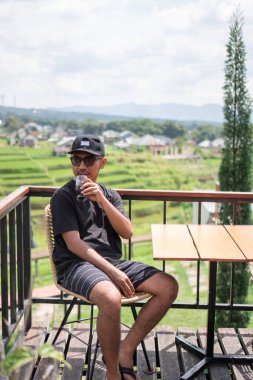 Siyah t-shirt ve şapka giyen Asyalı bir adam, Endonezya 'nın manzaralı dağ ve pirinç tarlalarının tadını çıkarırken terasta oturup kahve içiyor. Rahatlatıcı yaşam tarzı ve seyahat kavramı.