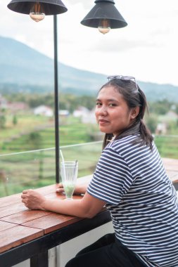 Asyalı bir kadın teras kafeteryasında oturmuş elinde taze bir içecek, çizgili gömlek ve saçında çiçek var. Endonezya 'daki pirinç tarlalarının ve dağların manzarası. Boş zaman, seyahat ve yaşam tarzı kavramı.