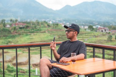 Siyah t-shirt ve şapka giyen Asyalı bir adam, Endonezya 'nın manzaralı dağ ve pirinç tarlalarının tadını çıkarırken terasta oturup kahve içiyor. Rahatlatıcı yaşam tarzı ve seyahat kavramı.