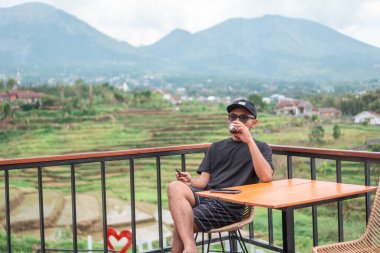 Siyah t-shirt ve şapka giyen Asyalı bir adam, Endonezya 'nın manzaralı dağ ve pirinç tarlalarının tadını çıkarırken terasta oturup kahve içiyor. Rahatlatıcı yaşam tarzı ve seyahat kavramı.