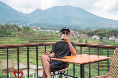 Siyah t-shirt ve şapka giyen Asyalı bir adam, Endonezya 'nın manzaralı dağ ve pirinç tarlalarının tadını çıkarırken terasta oturup kahve içiyor. Rahatlatıcı yaşam tarzı ve seyahat kavramı.