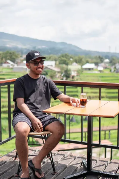 Siyah t-shirt ve şapka giyen Asyalı bir adam, Endonezya 'nın manzaralı dağ ve pirinç tarlalarının tadını çıkarırken terasta oturup kahve içiyor. Rahatlatıcı yaşam tarzı ve seyahat kavramı.