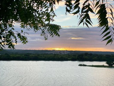 Sakin bir gün batımı, sakin bir göle yansıyan canlı renkler, üzerinde asılı ağaç dalları ile çerçevelenmiş, barışçıl ve resimsel bir manzara yaratır..