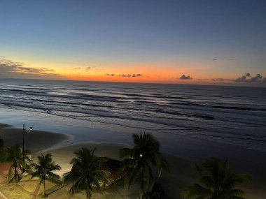 Sakin bir tropikal sahilde çarpıcı bir gündoğumu manzarası palmiye ağaçları, sakin okyanus dalgaları ve dinginlik ve huzuru temsil eden parlak turuncu ve mavi gökyüzü..