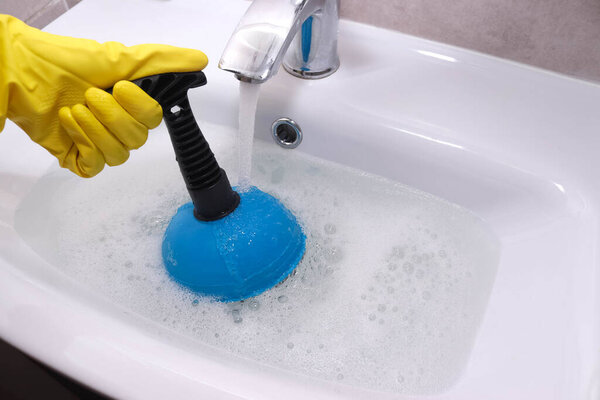 Clogged drain in bathroom sink. Hand in yellow gloves cleaning sink with plunger.