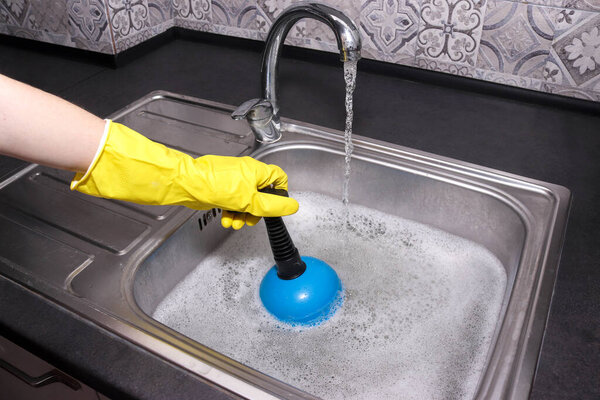 Hand in yellow gloves cleaning sink with plunger. Overflowing kitchen sink. Clogged drain.